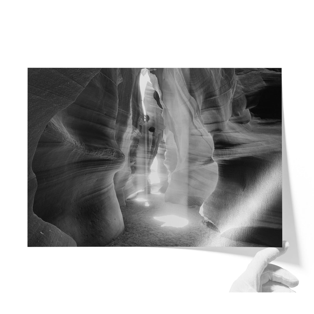 A black and white photograph of a narrow, winding path leading through a canyon or rock formation, with sunlight streaming through the opening and casting shadows on the ground.