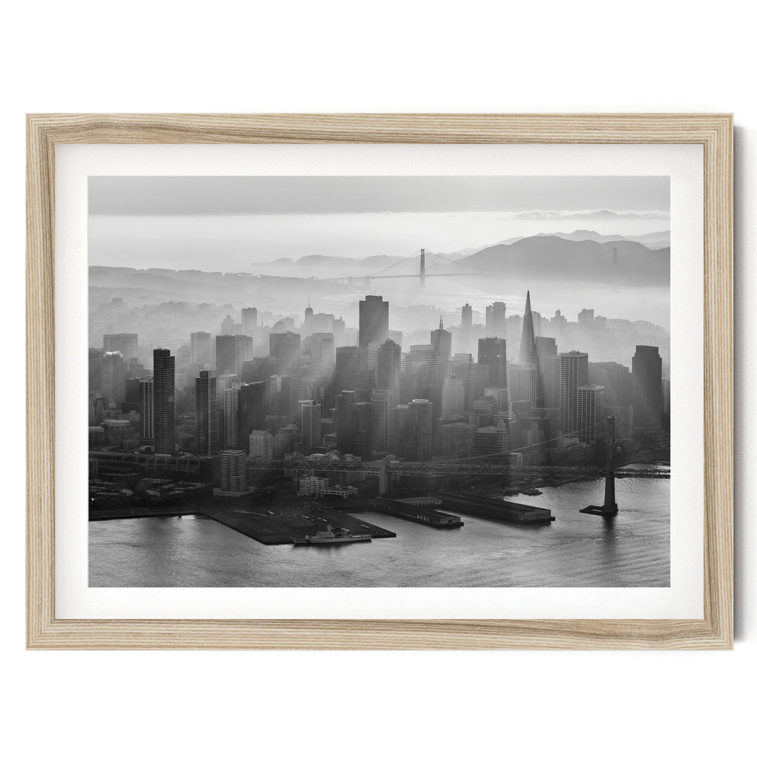 A black and white aerial view of a densely populated city skyline, with tall buildings and skyscrapers stretching out across the frame. The city is situated near a body of water, with a bridge visible in the distance. The sky above the city is hazy, creating a dramatic and atmospheric effect.