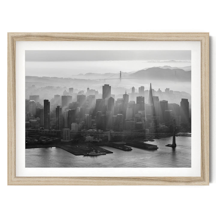 A black and white aerial view of a densely populated city skyline, with tall buildings and skyscrapers stretching out across the frame. The city is situated near a body of water, with a bridge visible in the distance. The sky above the city is hazy, creating a dramatic and atmospheric effect.