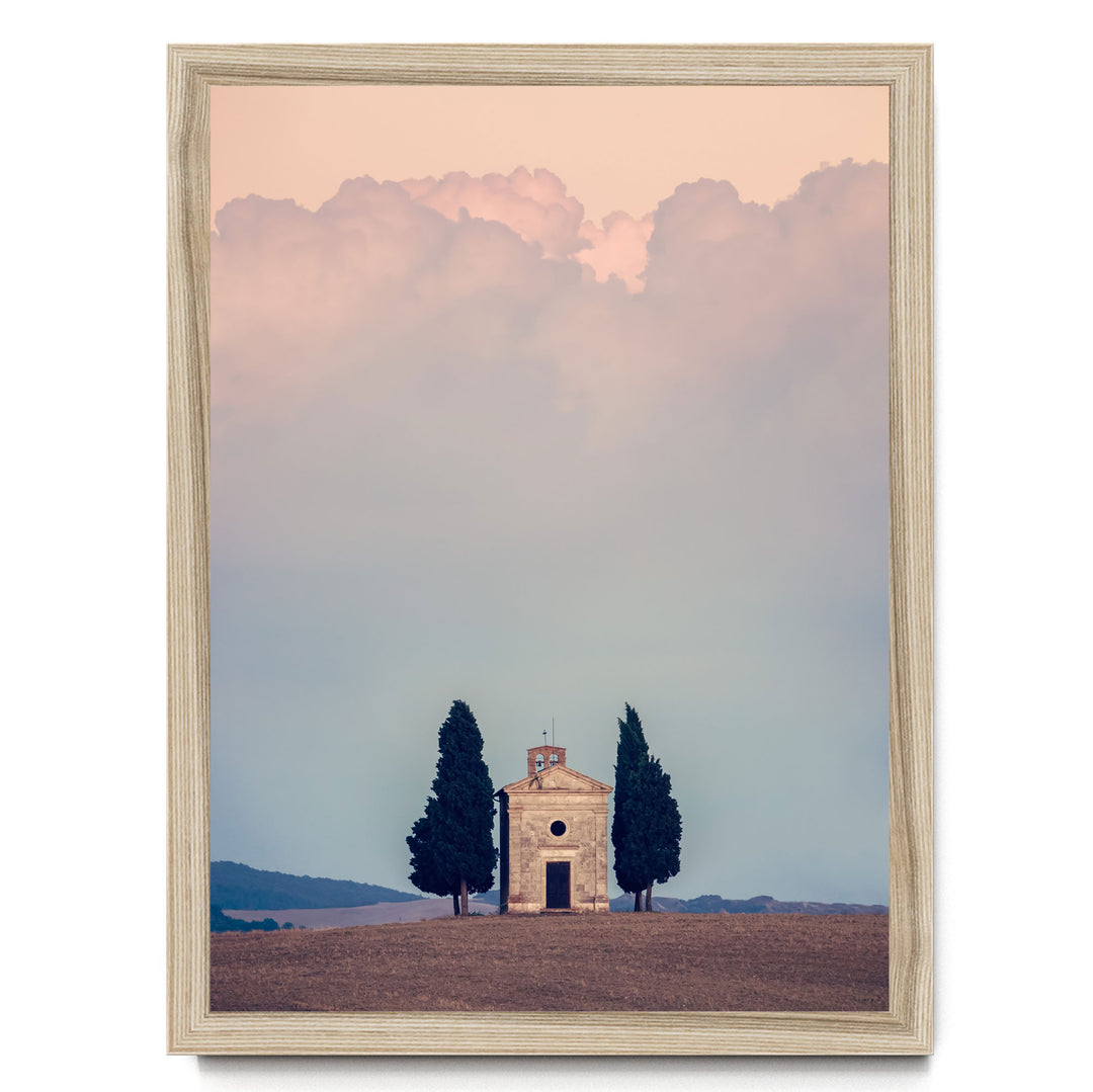 A small stone church with a bell tower sits in a field, surrounded by two tall cypress trees, with a cloudy sky in the background.
