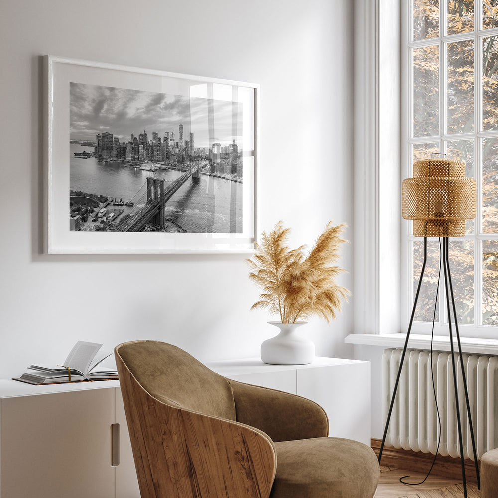a modern living room with a large framed black and white photograph of the Brooklyn Bridge hanging on the wall. The photograph captures the bridge and the city skyline, creating a striking focal point in the room. The room is furnished with a comfortable brown armchair, a white side table, and a floor lamp. A vase with dried flowers adds a touch of natural beauty to the space.