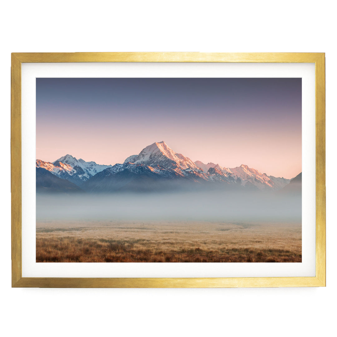 A framed landscape photograph depicting a mountain range with snow-capped peaks and a hazy, golden-hued sky at sunset.