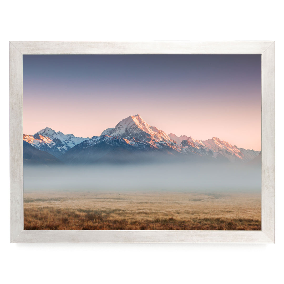 a breathtaking mountain range with snow-capped peaks, set against a vibrant sky at sunset. The mountains are partially shrouded in a hazy mist, creating a sense of depth and distance. The scene is framed within a white border, enhancing the overall composition and making it a visually striking image.