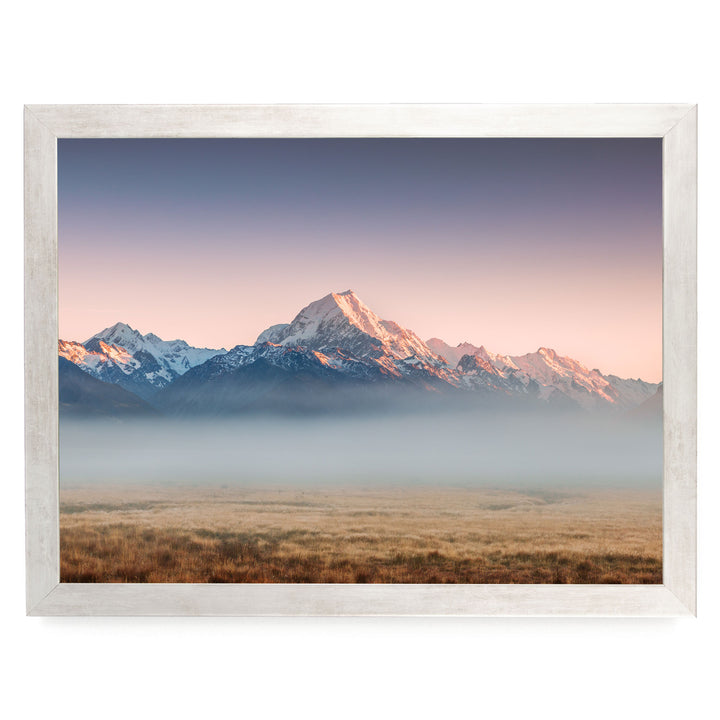 a breathtaking mountain range with snow-capped peaks, set against a vibrant sky at sunset. The mountains are partially shrouded in a hazy mist, creating a sense of depth and distance. The scene is framed within a white border, enhancing the overall composition and making it a visually striking image.