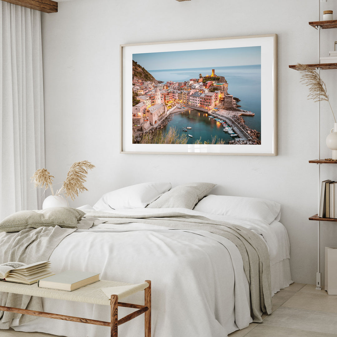 a bedroom with a large framed picture of a coastal town hanging on the wall above a bed with white linens and pillows.
