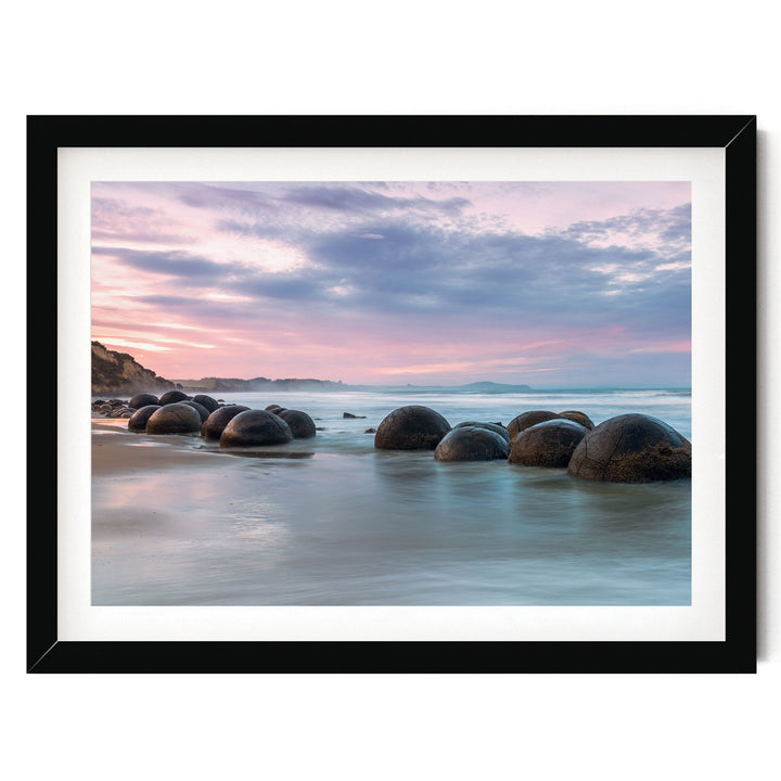 A serene beach scene with a collection of large, round boulders in the water, set against a picturesque sky with pink and blue hues.