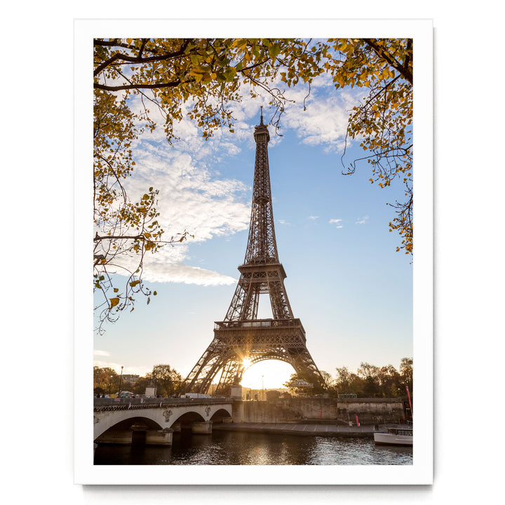 the Eiffel Tower in Paris, France, with a bridge and trees in the foreground, and the sun setting behind the tower.