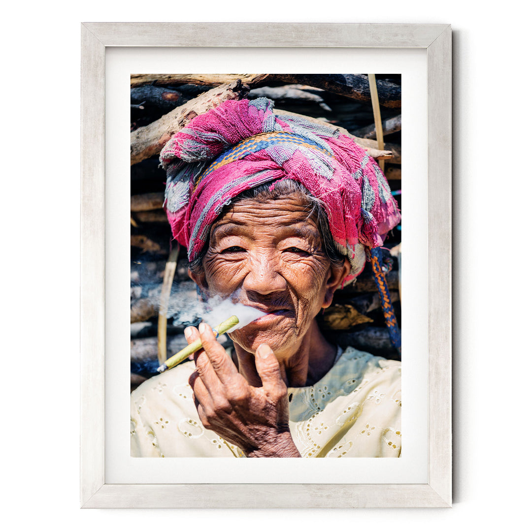 An elderly woman wearing a pink headscarf and a white blouse is shown smoking a cigarette, with a pile of wood visible in the background.