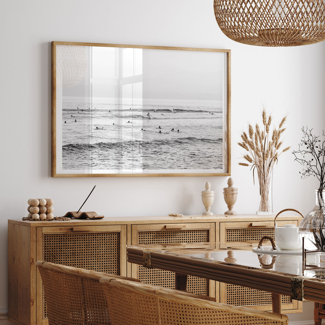 a serene beach scene with a framed photograph hanging on the wall, depicting people swimming in the ocean. The photograph is framed in a wooden frame and is placed above a wooden cabinet or dresser. The cabinet has a wicker design and is situated in a dining area, with a table and chairs visible in the foreground.