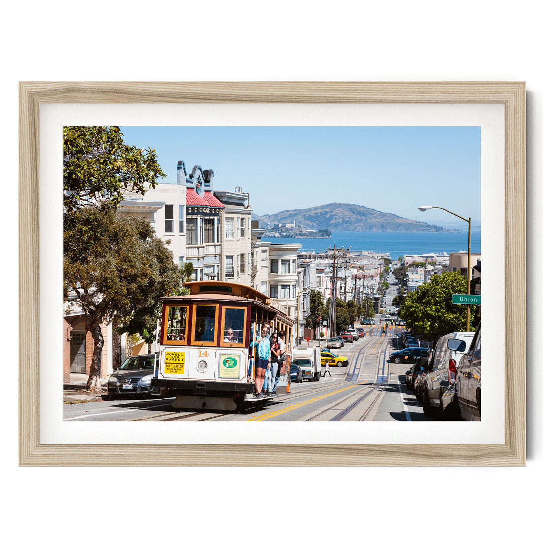 A cable car is traveling down a busy street in San Francisco, with buildings, cars, and pedestrians visible in the background.