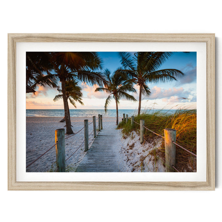 A wooden walkway leads to a beach lined with palm trees, with the ocean visible in the distance.