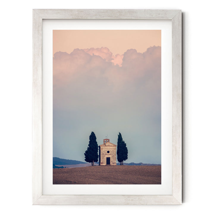 A framed photograph depicting a small white church with a bell tower, surrounded by two tall trees, set against a cloudy sky.