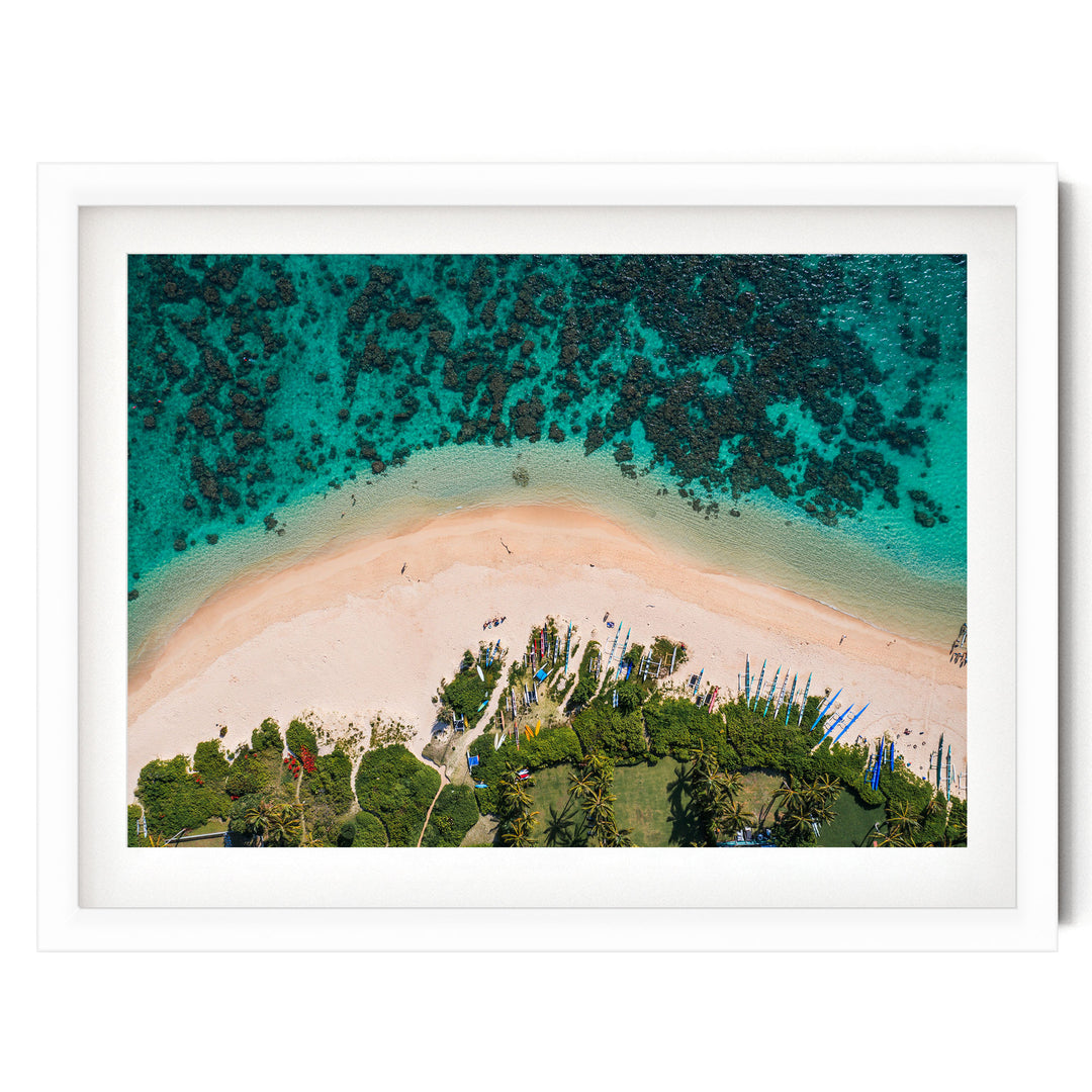 The image is an aerial view of a tropical beach with crystal clear turquoise waters, sandy shores, lush vegetation, and several boats on the water.
