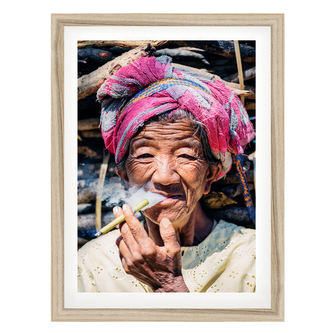 An elderly woman wearing a pink headscarf and a white blouse is shown smoking a cigarette, with a pile of wood visible in the background.