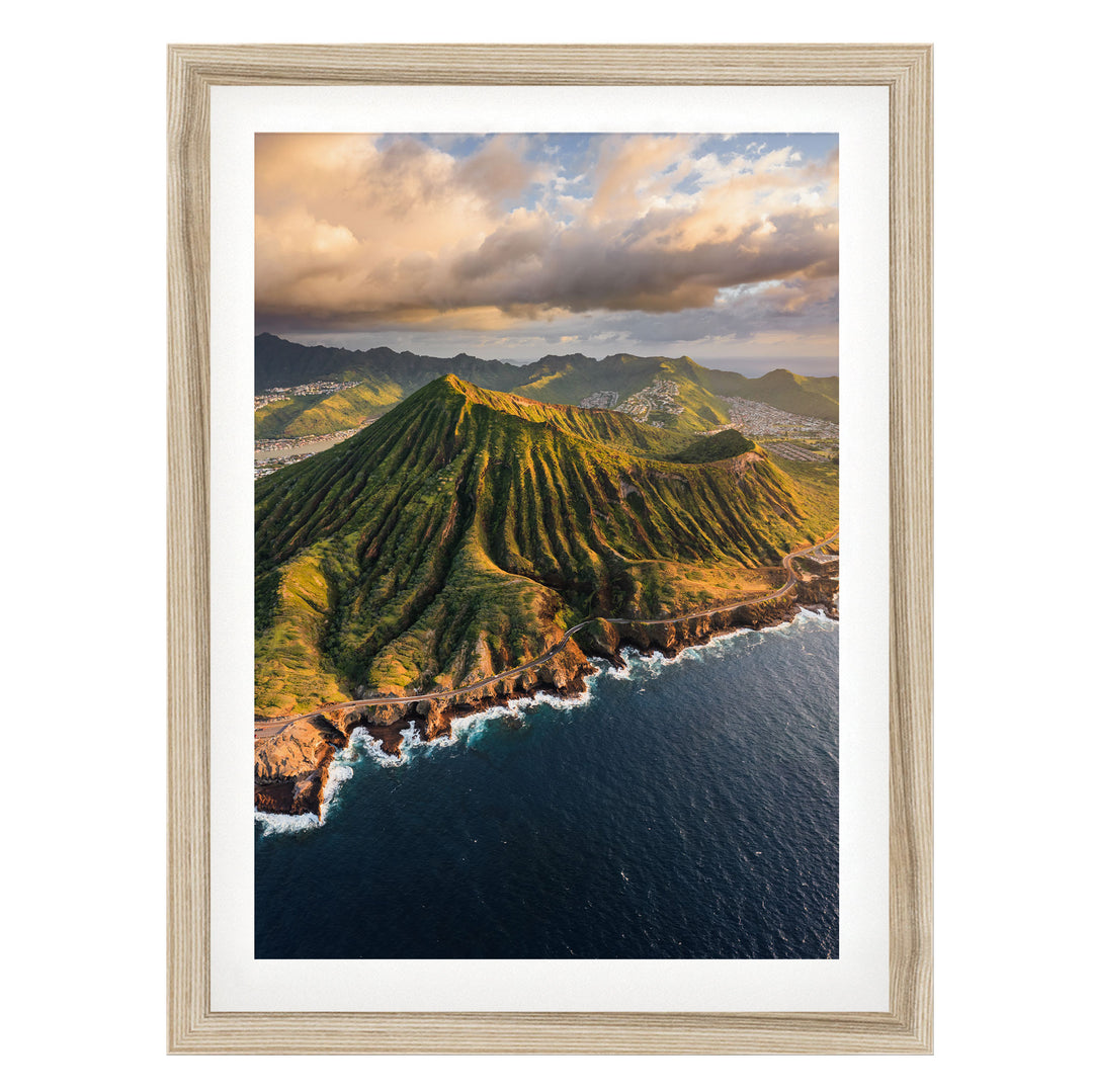 a mountainous coastline with a steep, jagged peak overlooking the ocean. The sky is filled with clouds, creating a dramatic backdrop for the scene. The photograph is framed and appears to be a high-quality image of a natural landscape.