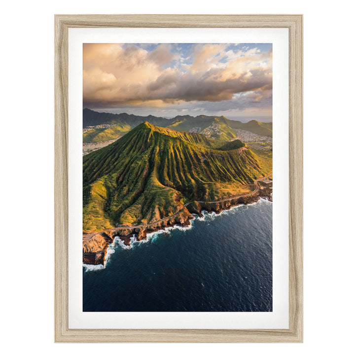 a mountainous coastline with a steep, jagged peak overlooking the ocean. The sky is filled with clouds, creating a dramatic backdrop for the scene. The photograph is framed and appears to be a high-quality image of a natural landscape.