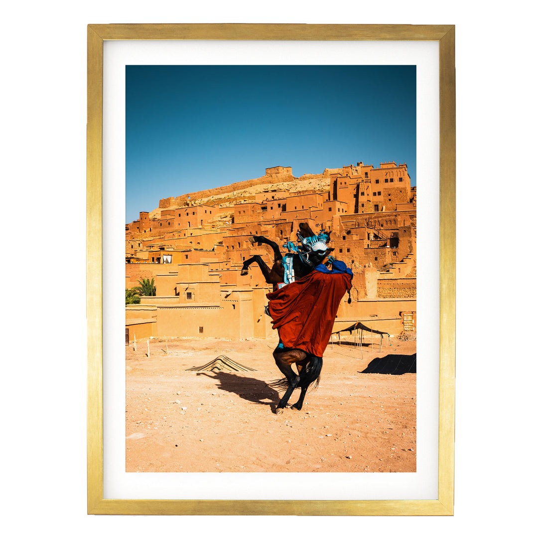 a person riding a rearing horse in a desert-like setting, with a large, multi-story structure in the background.