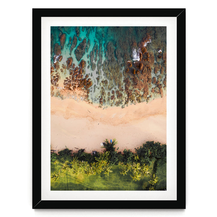The image is a framed aerial view of a beach with a mix of sandy and rocky areas, surrounded by lush greenery and palm trees.