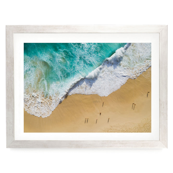 The image is a framed photograph of a beach scene, featuring a large wave crashing onto the sandy shore and people walking along the beach.