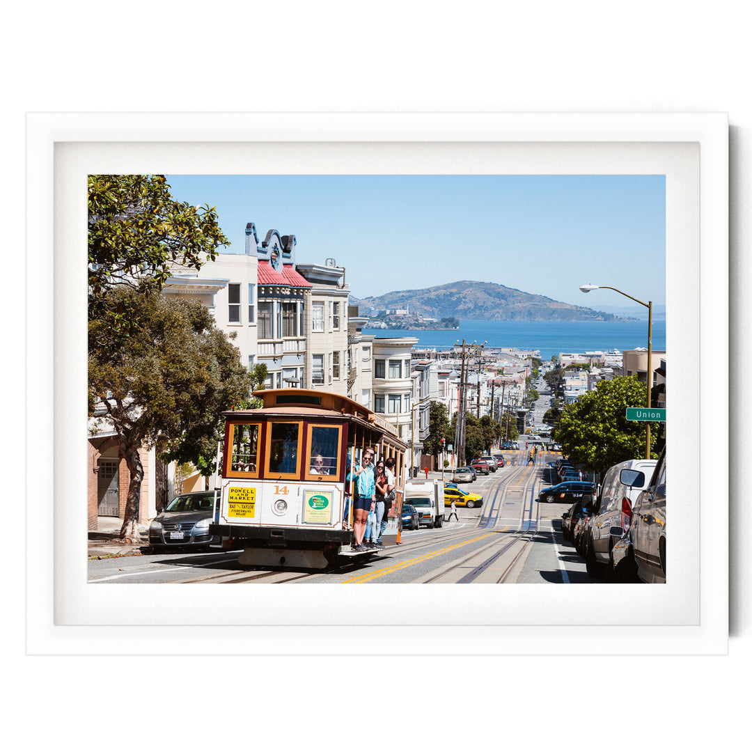 A cable car is traveling down a street lined with buildings and trees, with a mountain visible in the distance.
