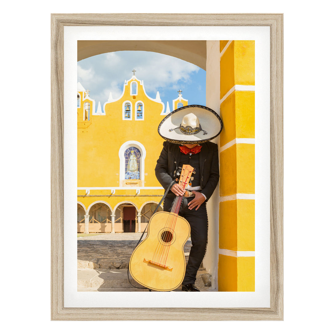 A person dressed in a traditional Mexican outfit, including a sombrero and a black suit, is holding a guitar and standing in front of a yellow building with arched windows.