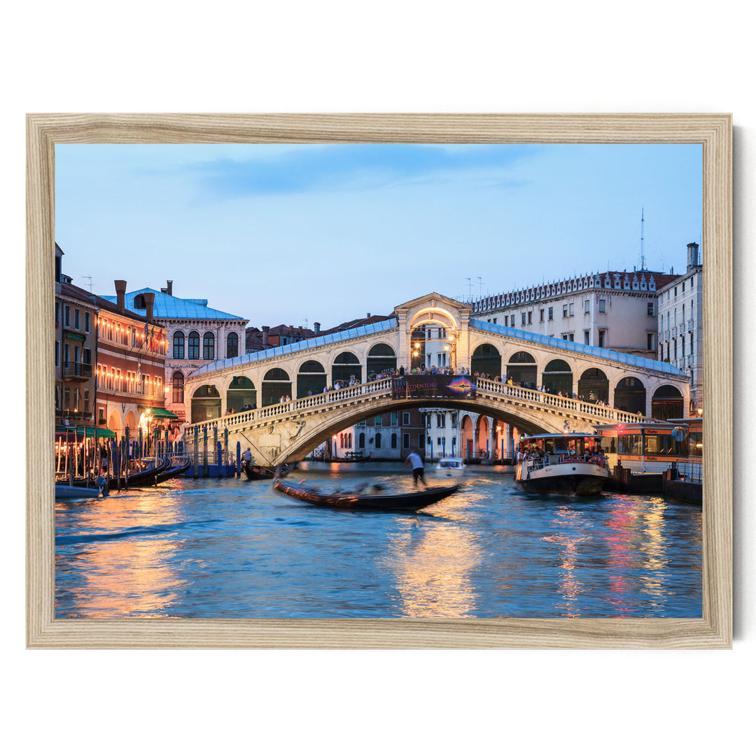 a picturesque scene of the Rialto Bridge in Venice, Italy, with its distinctive white stone arch spanning the Grand Canal. The bridge is illuminated by warm lights, creating a romantic atmosphere. The canal is filled with boats, adding to the lively ambiance of the scene. The image is framed, suggesting it is a piece of art or a photograph.
