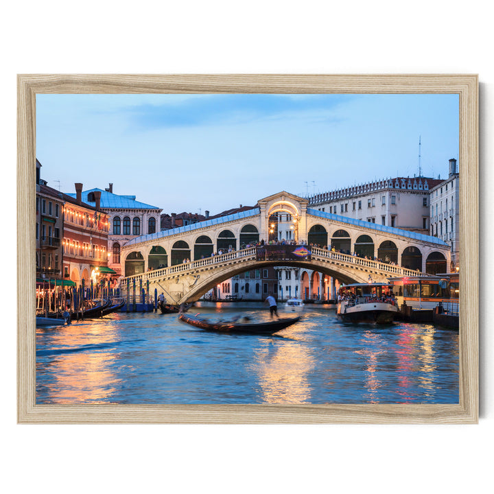 a picturesque scene of the Rialto Bridge in Venice, Italy, with its distinctive white stone arch spanning the Grand Canal. The bridge is illuminated by warm lights, creating a romantic atmosphere. The canal is filled with boats, adding to the lively ambiance of the scene. The image is framed, suggesting it is a piece of art or a photograph.