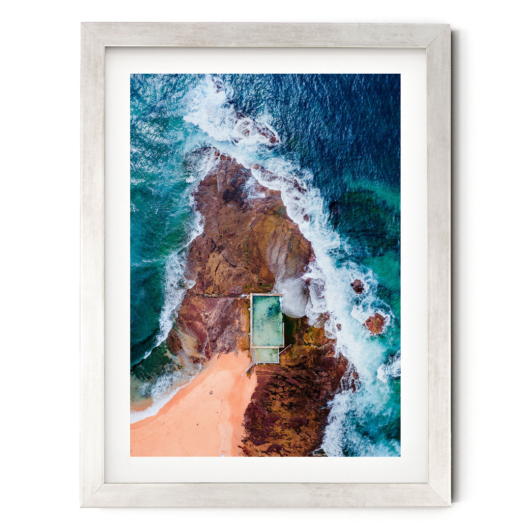 The image is an aerial view of a beach with a swimming pool, surrounded by rocky cliffs and crashing waves.