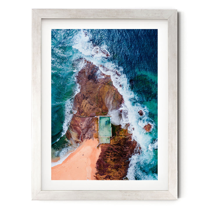 The image is an aerial view of a beach with a swimming pool, surrounded by rocky cliffs and crashing waves.
