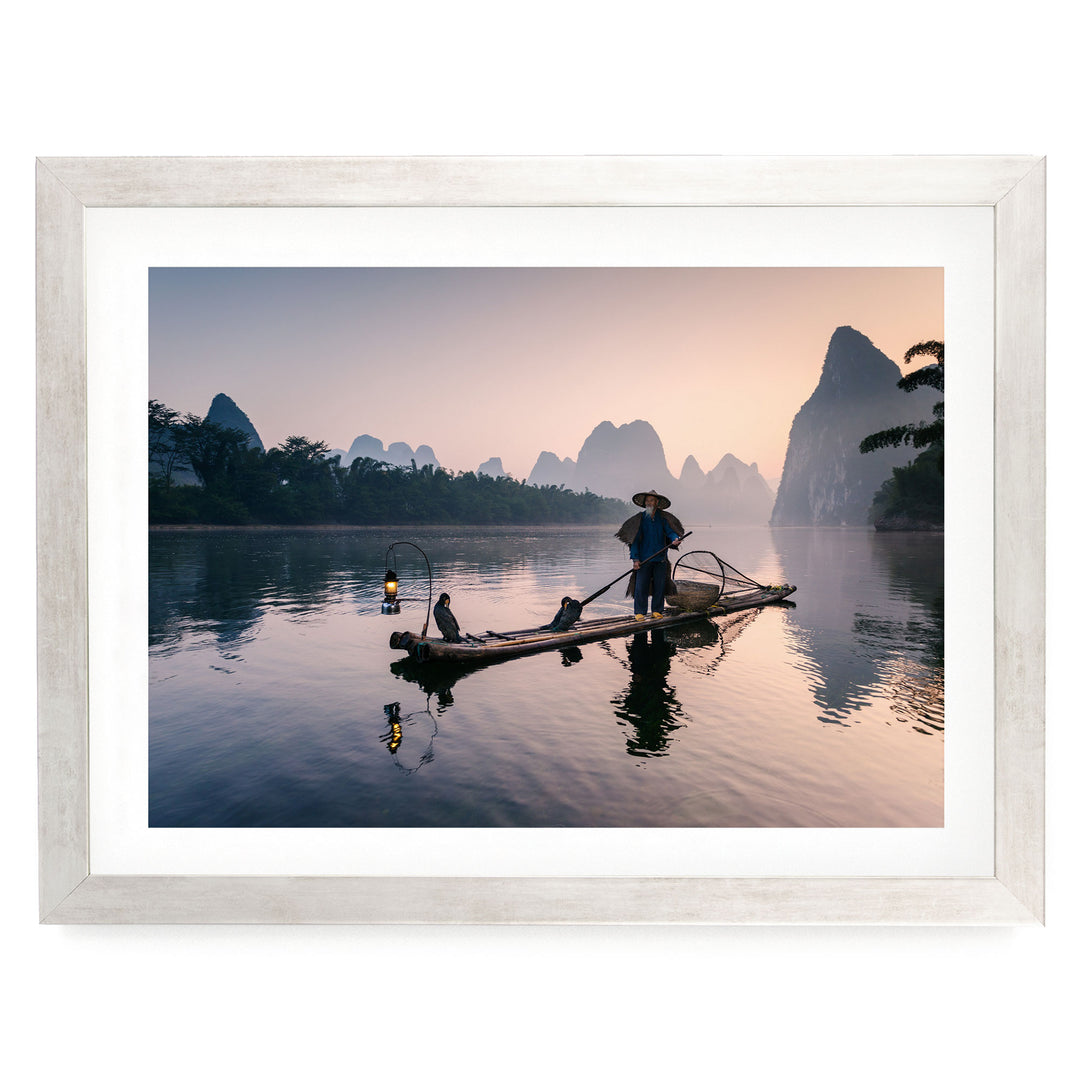 A person stands on a long wooden boat, paddling through a serene lake at sunset, with mountains in the background.
