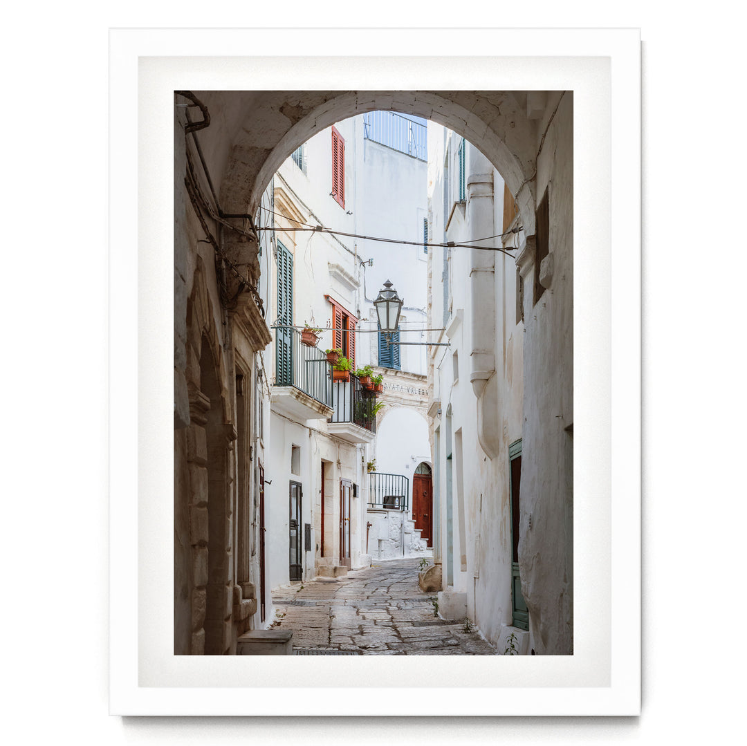 A narrow cobblestone street lined with white buildings and colorful shutters, leading to a balcony with potted plants.