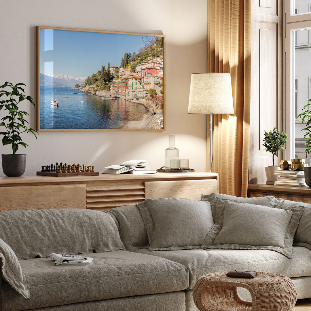 a cozy living room with a large framed picture of a coastal town hanging on the wall, a comfortable gray sofa, a wooden side table with a lamp, and various decorative elements such as potted plants and books.