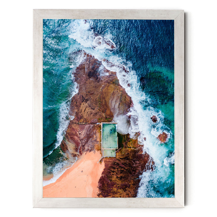 The image is an aerial view of a beach with a swimming pool, surrounded by rocky cliffs and crashing waves.