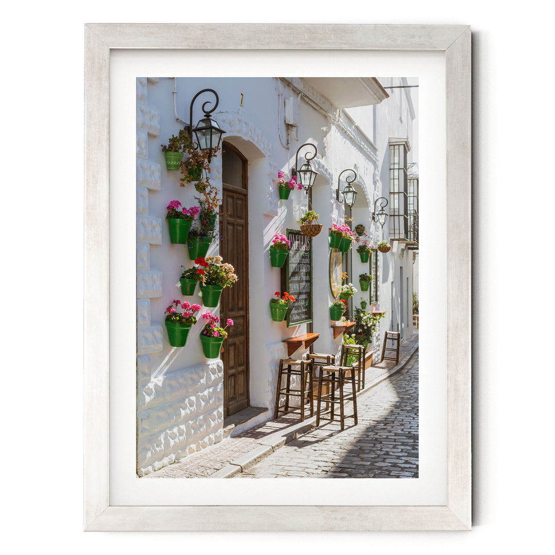 A charming street scene with a white building adorned with hanging flower pots and a wooden door. The building has a quaint, rustic charm, and the cobblestone street adds to the picturesque atmosphere.