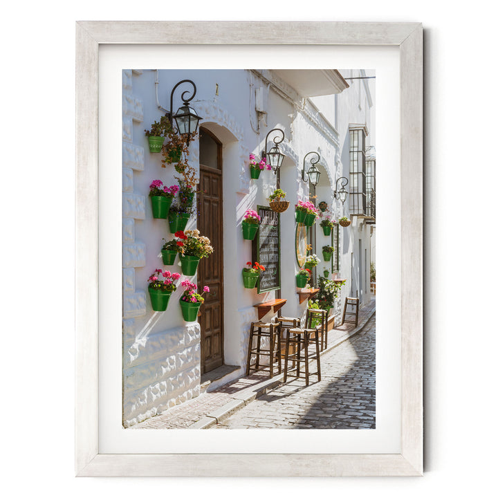 A charming street scene with a white building adorned with hanging flower pots and a wooden door. The building has a quaint, rustic charm, and the cobblestone street adds to the picturesque atmosphere.
