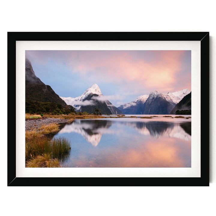 A framed photograph depicting a serene landscape with snow-capped mountains reflected in a calm lake, set against a vibrant sky with pink and orange hues.