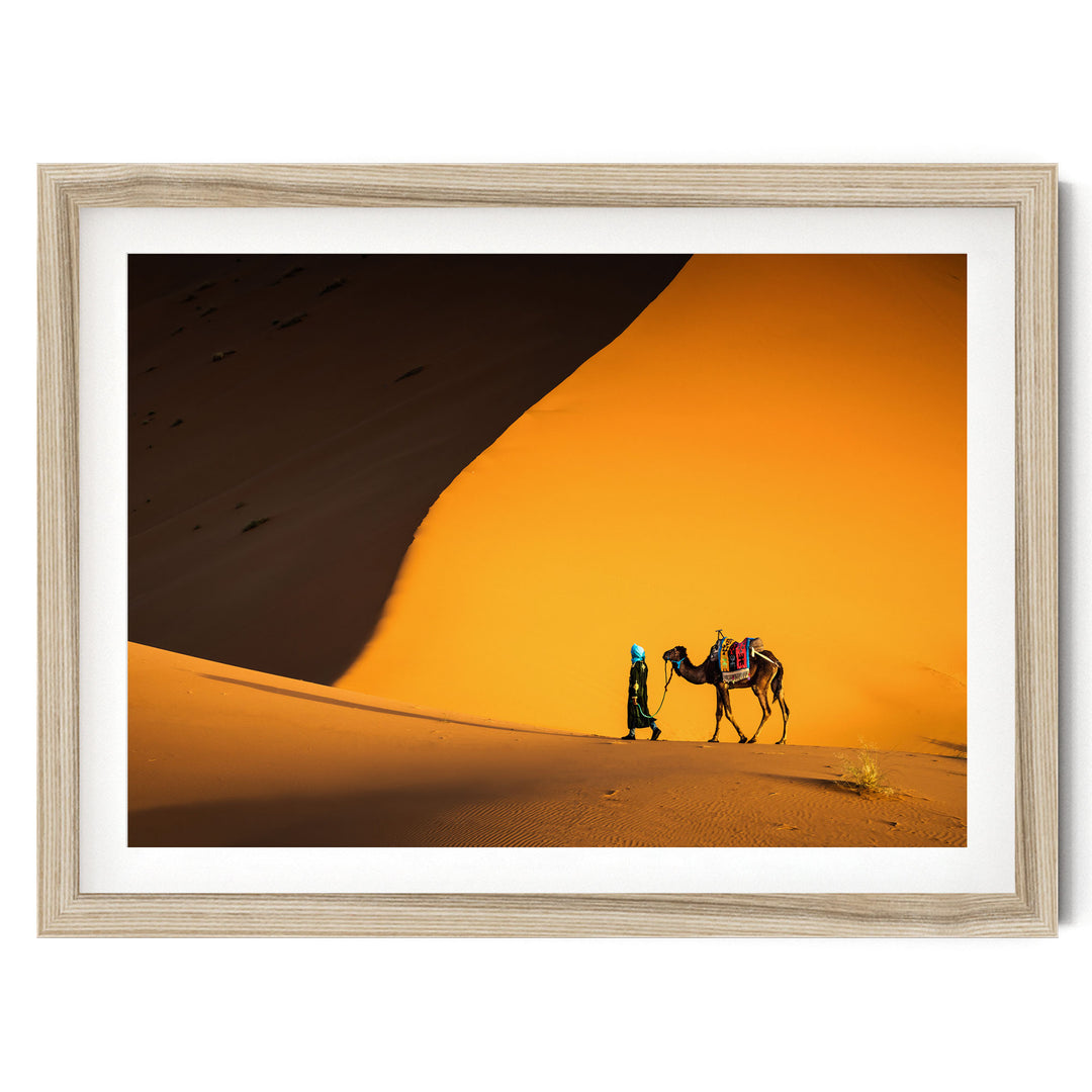 A person is walking a camel across a desert landscape with a large sand dune in the background.