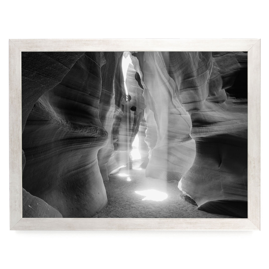A black and white photograph of a narrow passage in a rock formation, with sunlight streaming through the opening and casting shadows on the walls.