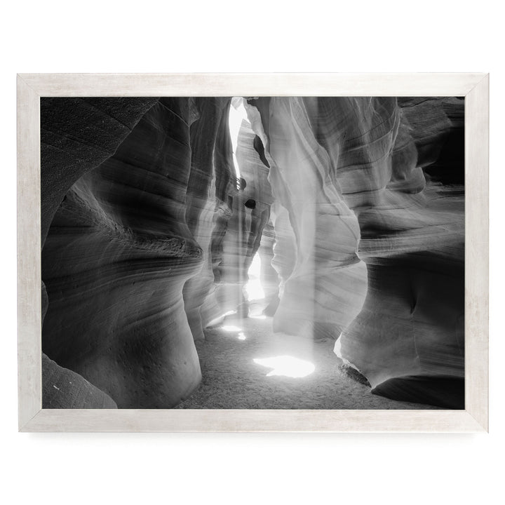 A black and white photograph of a narrow passage in a rock formation, with sunlight streaming through the opening and casting shadows on the walls.