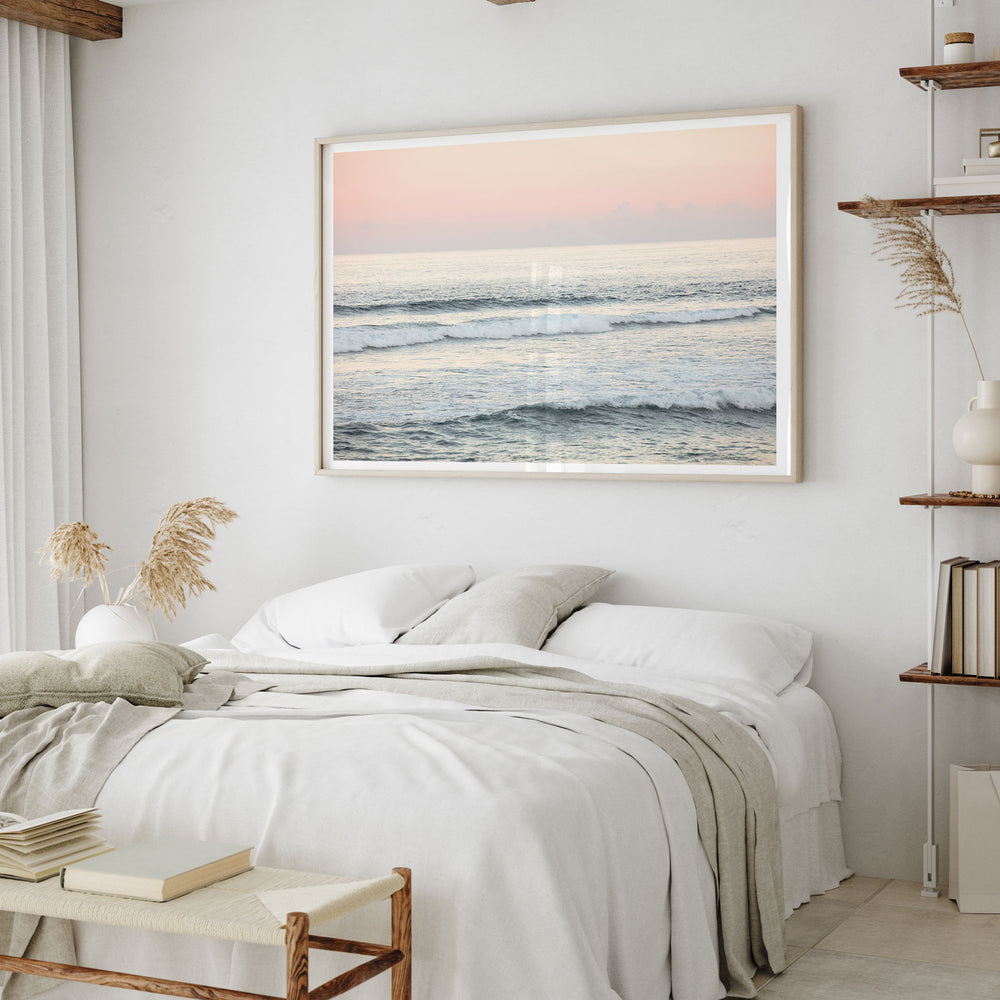 a serene bedroom scene with a large framed picture of a beach at sunset hanging on the wall above a bed with white linens and a gray throw blanket.