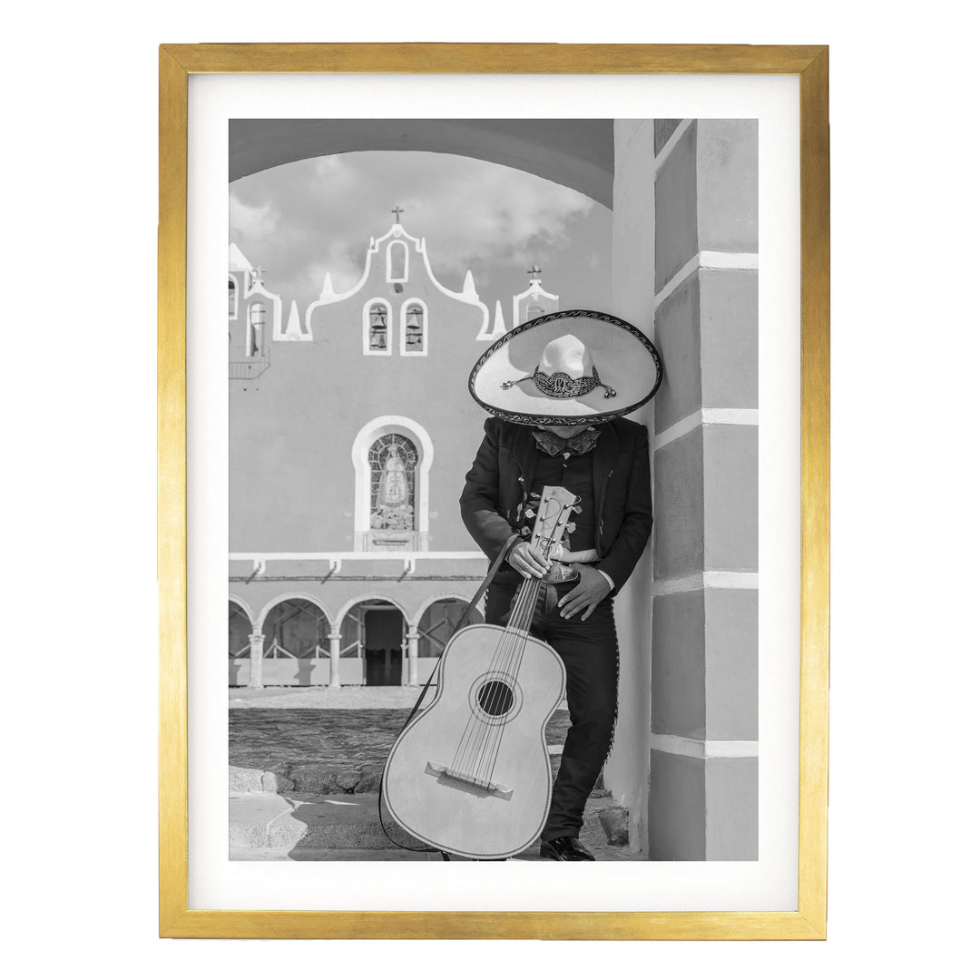 A person wearing a sombrero and holding a guitar stands in front of a building with arched windows and a cross on top.