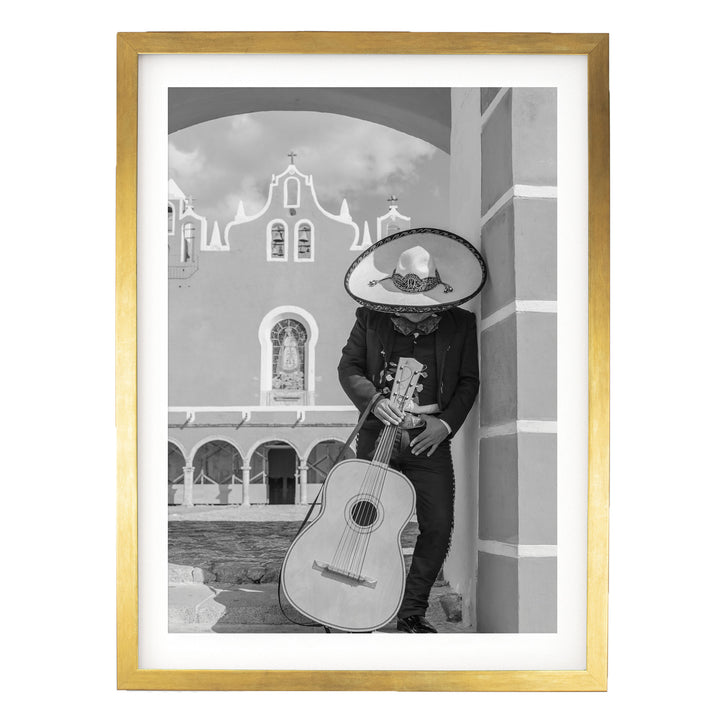 A person wearing a sombrero and holding a guitar stands in front of a building with arched windows and a cross on top.