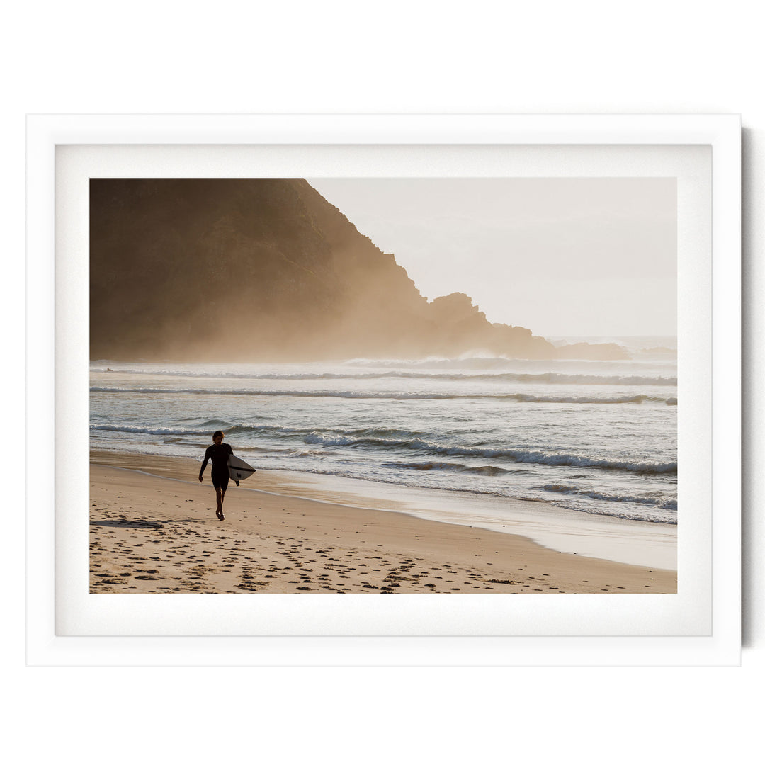 A surfer walks along the beach, carrying a surfboard, with a rocky cliff and misty mountains in the background.