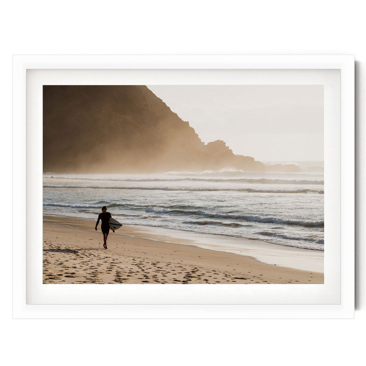 A surfer walks along the beach, carrying a surfboard, with a rocky cliff and misty mountains in the background.
