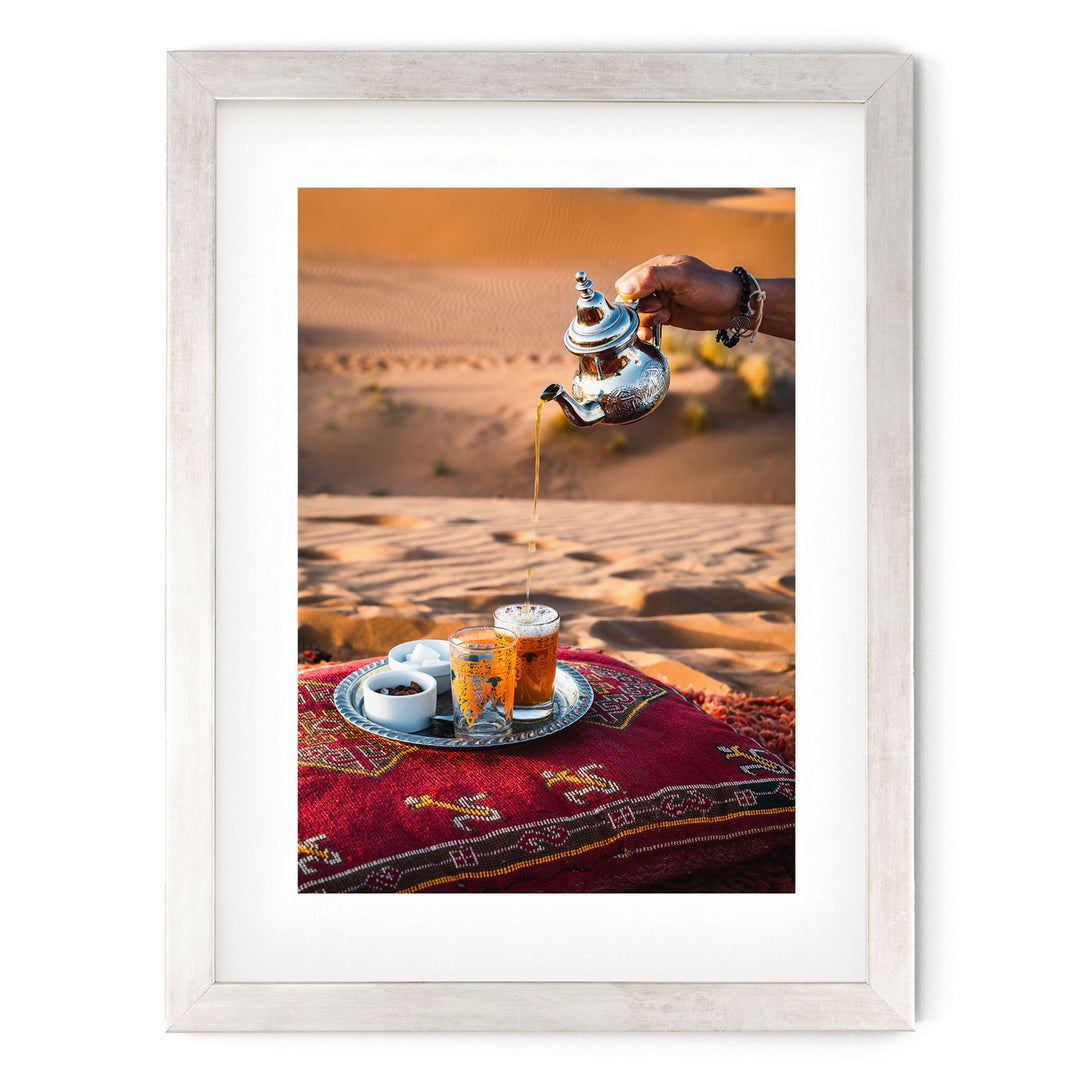 A person is pouring tea from a silver teapot into a glass, with a tray of tea and other items on a red and gold patterned rug.