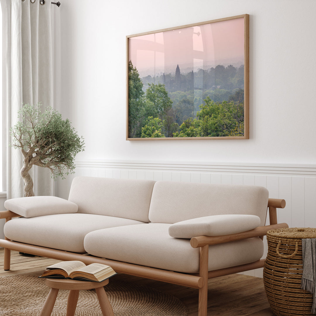 a cozy living room with a large framed landscape painting hanging on the wall above a beige sofa. The sofa is positioned next to a small table with a book on it. The room also includes a potted plant and a woven basket, adding to the overall aesthetic of the space.