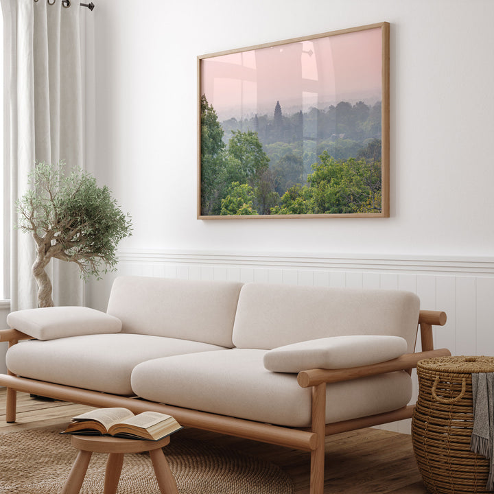a cozy living room with a large framed landscape painting hanging on the wall above a beige sofa. The sofa is positioned next to a small table with a book on it. The room also includes a potted plant and a woven basket, adding to the overall aesthetic of the space.