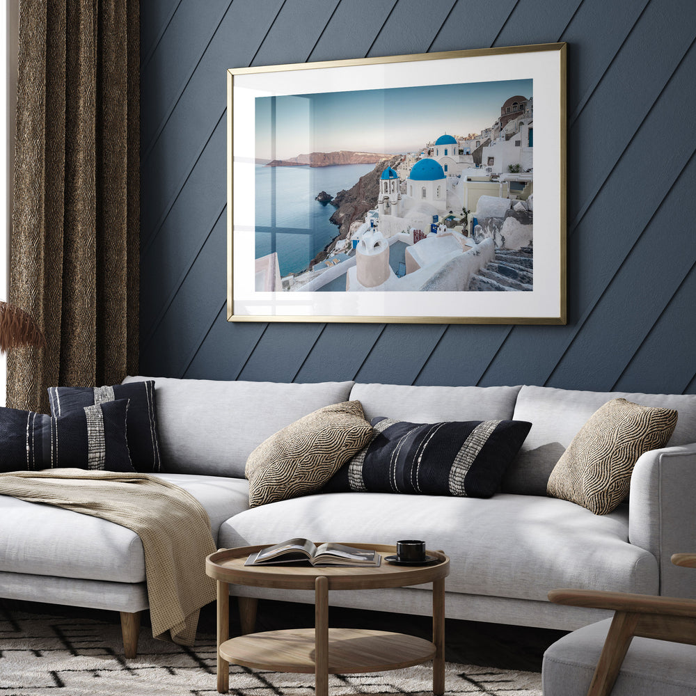 a living room with a large framed picture of a scenic view of a Greek island, specifically Santorini, hanging on the wall above a white sofa with patterned throw pillows.