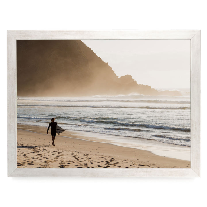 A person in a wetsuit carrying a surfboard walks along the beach, with a rocky cliff and ocean in the background.
