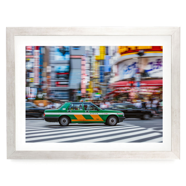 A green taxi cab is captured in motion, driving through a busy city street lined with colorful buildings and blurred pedestrians.